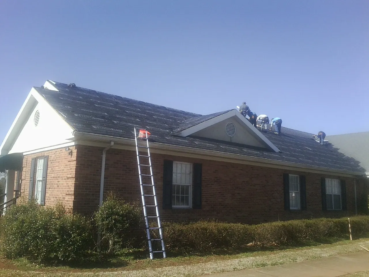 Crew tearing shingles off a brick ranch roof with a ladder set — Emergency Roof Repair work by our crew in Braggs