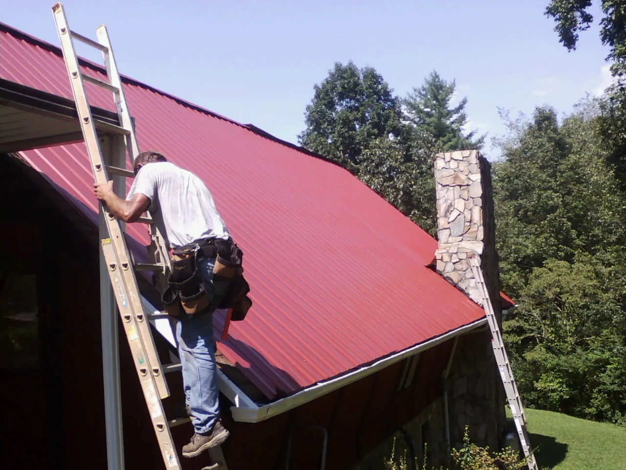 Roofer on a ladder beside a red metal roof and stone chimney — Soffit & Fascia Repair by Roofing Brothers in Braggs
